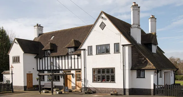 White rendered Arts and Crafts style pub with a steep brown tiled roof, tall white chimneys and dormers, a mock Tudor half-timbered centre section, mullioned windows and a timber sign frame at the front