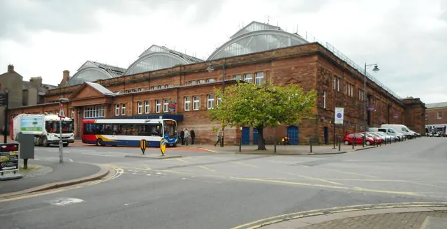 Large red sandstone Victorian market hall with long rows of windows and great arched glass roofs, a bus and parked vans along the frontage and a young tree on the pavement