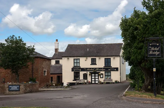 Cream rendered two-storey inn with a grey slate roof and a single chimney, a black fascia reading The Metal Bridge above a glazed double door with benches either side, a matching hanging sign on a post among trees to the right, and a low red sandstone barn and a car park sign to the left