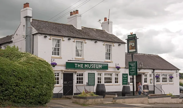 Two-storey whitewashed pub with a slate roof and tall chimneys, a long green sign reading The Museum across the front, barrels by the door, and a Greene King post sign beside a lower single-storey wing, with a large hedge in the foreground