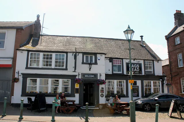 Two-storey whitewashed pub with black window frames and a slate roof, a sign reading Nags Head on the upper floor and Welcome above the door, picnic tables with people sitting outside on a cobbled square and a green lamp post in front