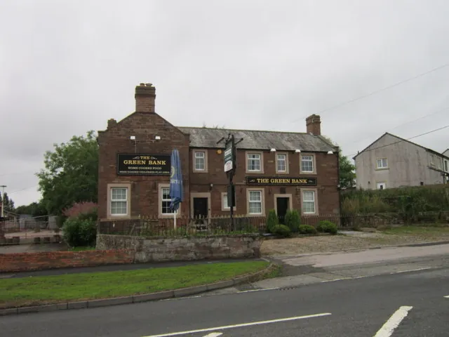 Red sandstone two-storey pub with a slate roof, end chimneys and cream window surrounds, black signs reading The Green Bank on the gable and over the doorways, a blue parasol and a pole sign out front, behind a low stone wall and grass verge on an overcast day