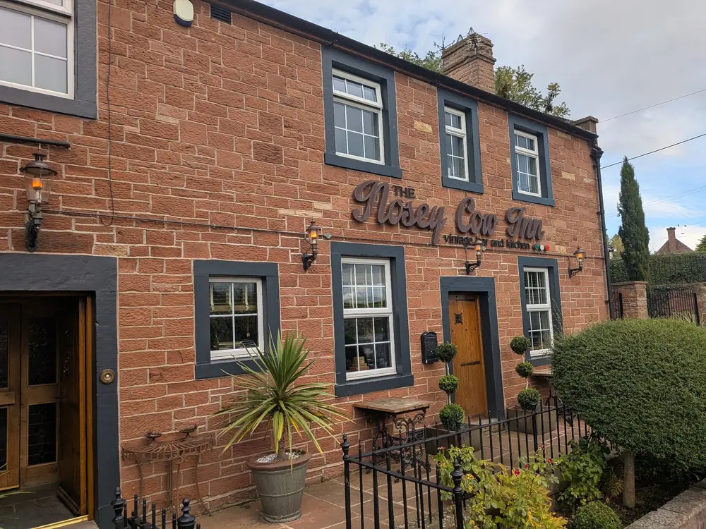 Red sandstone two-storey inn with dark grey window surrounds, the words The Nosey Cow Inn, vintage bar and kitchen in raised lettering between the floors, iron lanterns on the wall, a wooden door, potted plants and a clipped hedge behind black railings