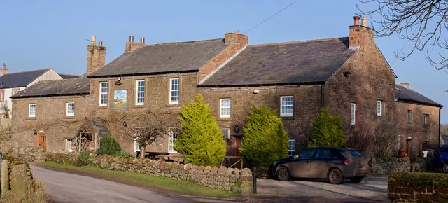 Long two-storey red sandstone village inn with slate roofs and several chimneys, a pictorial sign on the front wall, conifers and a stone wall along the front and a car parked at the side