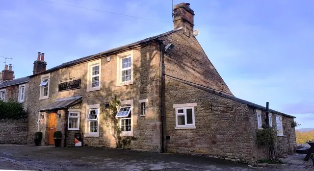 Two-storey sandstone village inn with a slate roof and white sash windows, a black sign reading The Plough Inn above a small porch and orange door, a lower stone wing stepping down to the right, and a climbing plant on the front wall