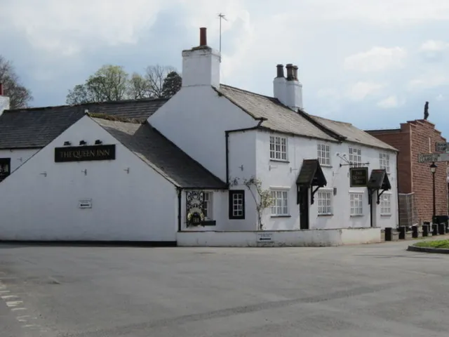 Whitewashed village pub of one and two storeys with grey slate roofs, white chimneys and small paned windows, a black sign reading The Queen Inn on the gable end, timber gabled porches over the doors and a hanging sign, facing an empty open green