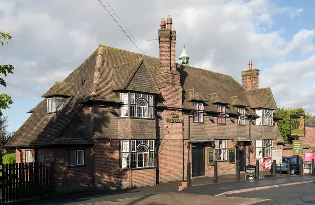 Large Arts and Crafts style brick pub with a steep tiled roof, several gabled dormers with bay windows, tall chimneys and a small green copper cupola, the name The Redfern lettered on the central chimney breast and a hanging pub sign on the right