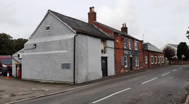 Roadside pub seen along a village street, with a plain grey rendered gable wing in the foreground bearing a Car Park to Rear sign, and beyond it a red brick frontage with white sash windows and a dark fascia sign reading The Royal Oak. A lower red brick building continues along the road.