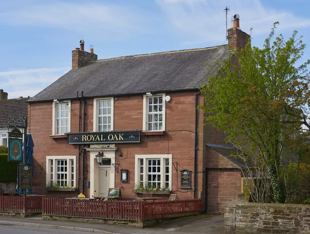 Two-storey red sandstone pub with a slate roof, white sash windows and a black fascia reading Royal Oak above a white doorway, a hanging sign on a post to the left, a red picket fence in front and a leafy tree to the right