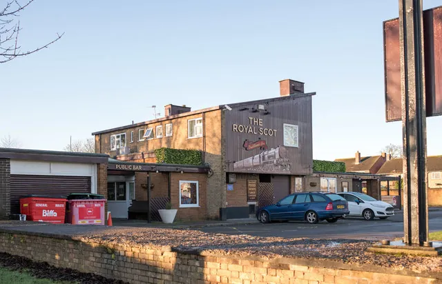 Flat-roofed brick estate pub with a tall dark timber clad end wall reading The Royal Scot above a painted steam locomotive, a low wing with a Public Bar sign over the entrance, and cars parked in front