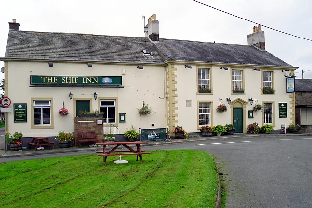 Long two-storey cream painted inn with a slate roof and stone quoins, a green fascia reading The Ship Inn, green doors, hanging baskets and window boxes, and a picnic bench on the grass in front