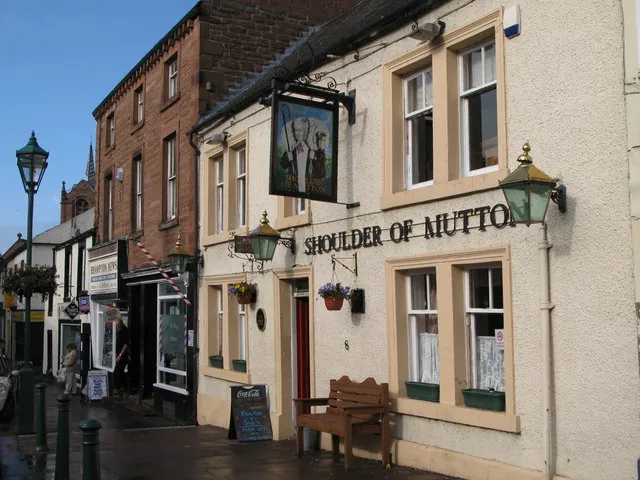 Cream rendered two-storey pub on a narrow street with stone window surrounds, black letters reading Shoulder of Mutton across the front, ornate green lanterns, hanging flower baskets, a wooden bench beneath the windows and a painted hanging sign of a figure, with shops and a church spire further along