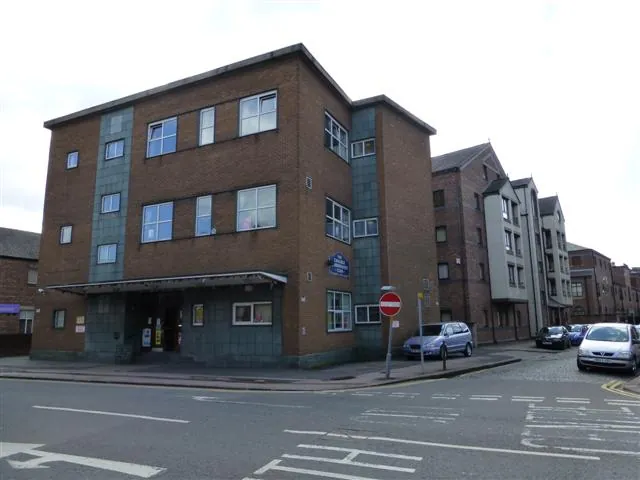 Modern four-storey red brick block with bands of blue grey tiles, a flat canopy over a glazed ground-floor entrance and a small round sign on the wall, on a street corner with a no entry sign and parked cars
