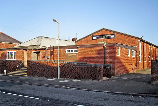 Single-storey red brick club building with a pitched gable end bearing a small sign reading St Margaret Mary's Social Club, a lower wing with a grey metal roof, and a low dark brick wall with steps along the pavement