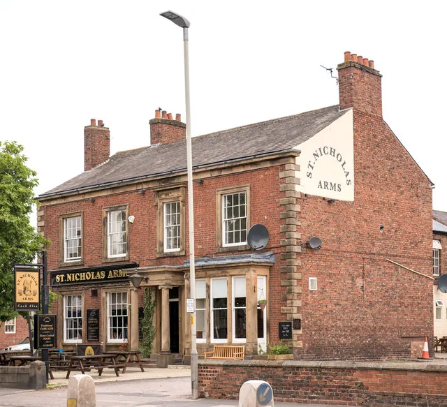 Red brick two-storey pub with sandstone quoins and window surrounds, a slate roof with chimneys, a black fascia reading St Nicholas Arms above the ground floor and the same name painted in a white panel on the gable end, with picnic tables and a hanging sign by the door