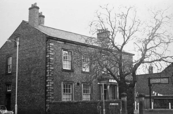 Black and white photograph of a plain two-storey brick house with stone quoins and a columned porch half hidden by a bare tree, a Grey Street sign on the boundary wall and a hanging pub sign on a post