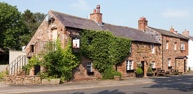 Long low two-storey pub of red sandstone with a slate roof and brick chimneys, its front largely covered in thick green ivy. An external stone stair with railings climbs the left gable, a hanging sign projects from the wall, and picnic benches stand by a row of cottages to the right in evening sun.