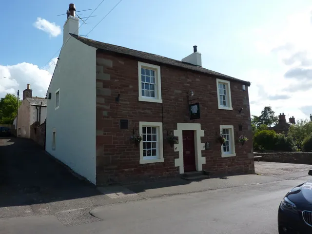 Two-storey red sandstone village inn with a whitewashed gable end, white sash windows, a dark red door in a pale stone surround, hanging baskets and a small hanging sign on the front wall, beside a lane
