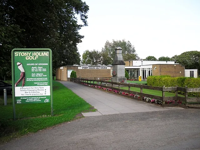 Low flat-roofed brick clubhouse with a glazed front seen at the end of a paved path lined with a timber fence and flower beds, a stone obelisk beside it, and a large green Stony Holme Golf noticeboard with opening hours in the foreground