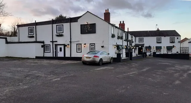 Long whitewashed two-storey inn with black trimmed eaves, a red brick chimney, lit wall lanterns and window boxes, a black panel with a white horse emblem on the front and a further white wing behind, seen across a large gravel car park at dusk with one silver car parked