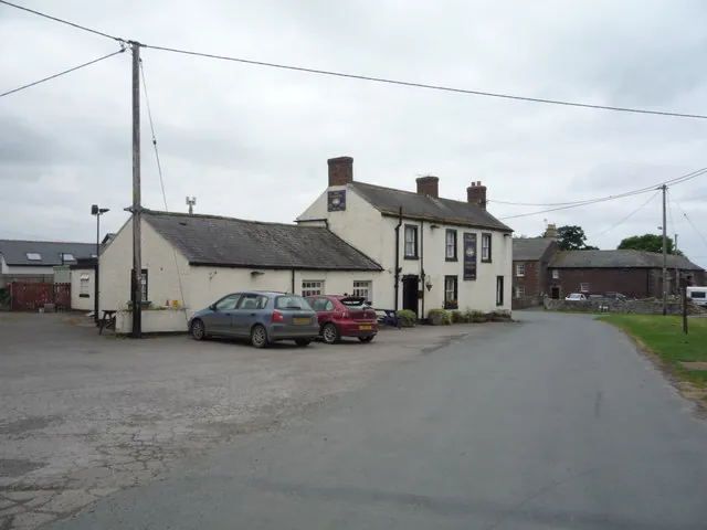 Small white rendered two-storey village pub with a slate roof and three chimneys, a dark hanging sign and a panel sign on the front, and a long single-storey whitewashed wing to the left, with two cars parked in front and a country road with telegraph poles under a grey sky