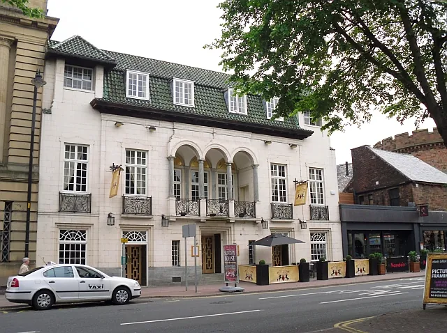 Cream faced two-storey building with a green tiled mansard roof and dormer windows, a recessed first-floor loggia of three round arches over the central doorway, wrought iron balconies at the first-floor windows and mustard coloured hanging banners, with planters and screens on the pavement and a white taxi parked in front