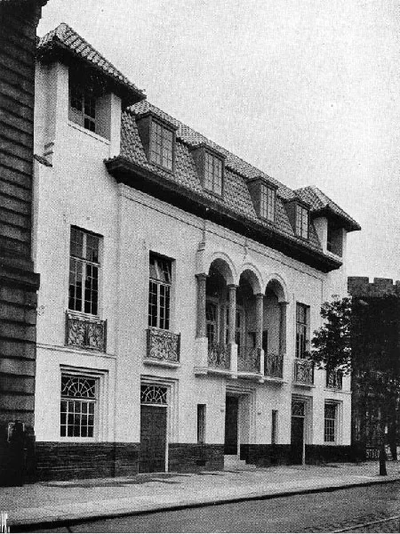 Black and white photograph of a pale rendered three-storey building with a steep tiled mansard roof and dormer windows, and a first-floor balcony of three round arches over a doorway, with ironwork balconies on the windows either side, a dark plinth, and a castellated stone wall at the right edge