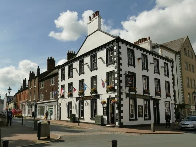 Three-storey white painted corner building with black window surrounds and black quoins, a gable with a tall chimney stack, window boxes of flowers and small flags along the first floor, and black fascia signs reading The Boardroom on both frontages, beside a row of red brick terraced houses