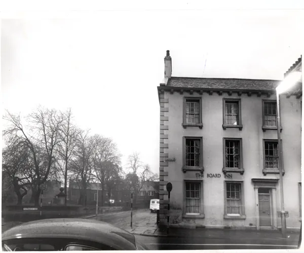 Black and white photograph of a tall three-storey rendered corner building with sash windows and stone quoins, the words The Board Inn on the front, bare trees across the road and the bonnet of a car in the foreground