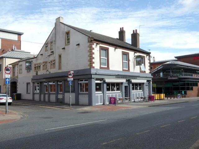 Two-storey corner pub with a white painted upper floor, brick window surrounds and a slate roof, and a grey painted ground floor with a row of windows and a few tables on the pavement, on a quiet street corner with modern buildings behind