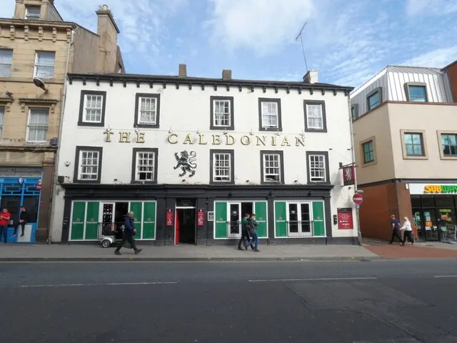 Three-storey white painted building with black window surrounds and gold lettering reading The Caledonian across the front, with a black heraldic lion between the words. The ground floor has a black shopfront with green shutters, and people walk past on the pavement of a town street.