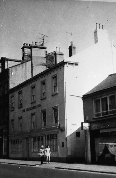 Black and white photograph of a three-storey rendered pub with plain sash windows and lettering reading The Caledonian Inn above large ground-floor windows, with two children walking past on the pavement