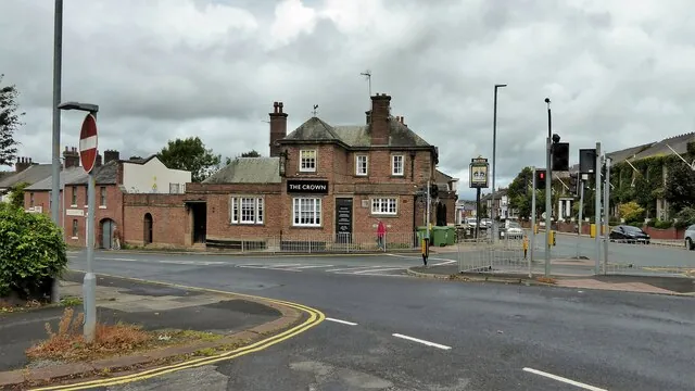 Red brick two-storey corner pub with a hipped slate roof, tall chimneys and a lower single-storey wing, a black sign reading The Crown over the ground-floor windows, seen across a wide road junction with traffic lights under a grey sky