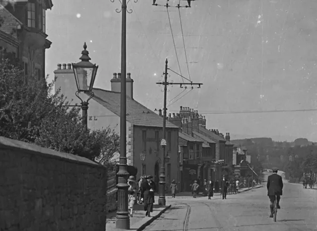 Black and white detail of a street with tram wires, a tall gas lamp in front of a white two-storey gabled building on the corner, a terrace running downhill beyond, and people on the pavement with a cyclist in the road