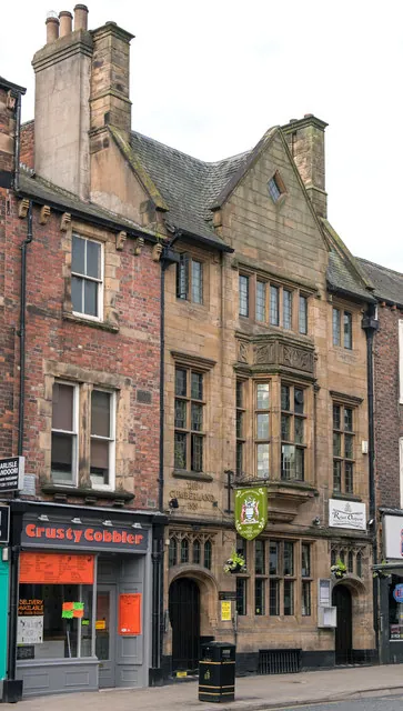 Tall three-storey sandstone building in a Tudor Gothic style, with a steep gable, mullioned windows, a carved oriel window and a green hanging sign over arched doorways, squeezed between a red brick building with a Crusty Cobbler shop and a narrower brick neighbour