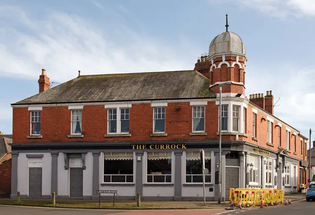 Two-storey red brick corner pub with a slate roof and a domed corner turret, white upper windows and a pale painted ground floor with grey pilasters, the name The Currock in gold letters on the fascia. Yellow roadwork barriers stand on the pavement at the corner.