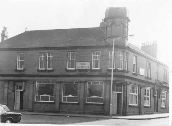Black and white photograph of a two-storey brick corner pub with a domed corner turret, a board reading The Currock between the upper windows, large ground-floor windows and a 1960s car parked at the left