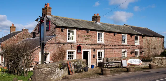 Long low two-storey red brick and sandstone pub with a grey slate roof and two chimneys, white framed windows and the words Drovers Rest lettered on the wall, a pole sign and a stone wall in front, and a banner on the forecourt reading National Pub of the Year