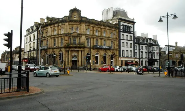 Ornate three-storey Victorian sandstone building with a clock in its central pediment and a balustraded roofline, a long sign reading The Griffin across the first floor, standing on a corner across a wide road junction with traffic lights and parked cars