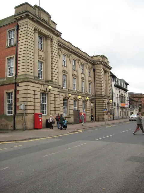 Large three-storey classical building of pale sandstone with tall rows of sash windows, pilasters and a heavy cornice, set beside an older red brick wing. Globe lamps on brackets line the ground floor, and a red pillar box and a few pedestrians stand on the pavement of a wide street.
