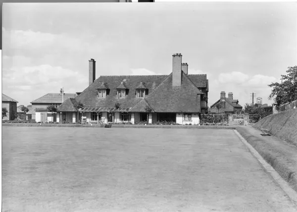 Black and white photograph across a level bowling green towards a long low roughcast building with a steep tiled roof, dormer windows and tall chimneys, a bench at the edge of the green