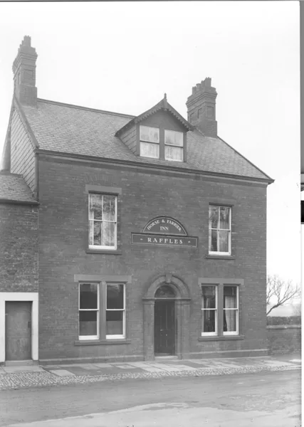 Black and white photograph of a narrow three-storey red brick house with a dormer gable, an arched board over the windows reading Horse and Farrier Inn and a sign beneath it reading Raffles