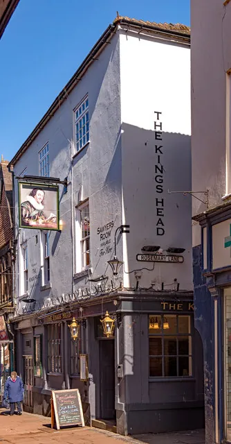 Tall narrow three-storey building with grey painted walls and a white rendered end wall bearing the words The Kings Head in tall black letters running down it, above a Rosemary Lane street sign. A painted hanging sign shows a bearded man in a ruff and wide hat, and brass lanterns and a chalkboard stand beside the dark ground-floor frontage on a narrow paved lane.