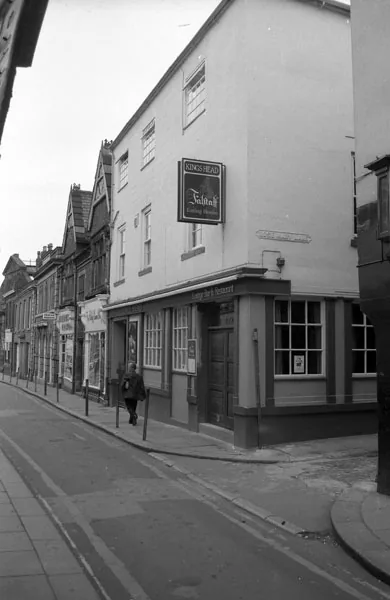 Black and white photograph looking along a narrow street to a three-storey rendered corner pub with a hanging sign reading Kings Head, a man walking past the door and older shopfronts beyond