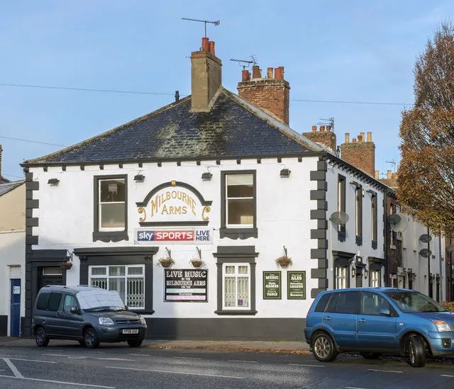 White painted two-storey corner pub with a slate roof, brick chimneys and dark quoins, the name Milbourne Arms in gold letters under a curved pediment above a Sky Sports Live sign. Boards advertise live music, hanging baskets hang between the windows and cars are parked outside.