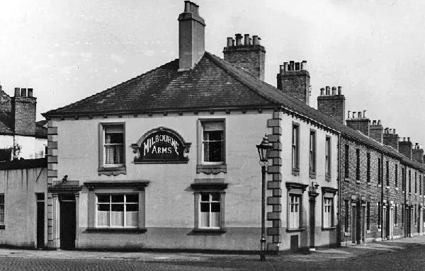 Black and white photograph of a two-storey rendered corner pub with stone quoins, an arched sign reading Milbourne Arms over the ground-floor window, a gas lamp at the corner and a terrace of brick houses running away to the right