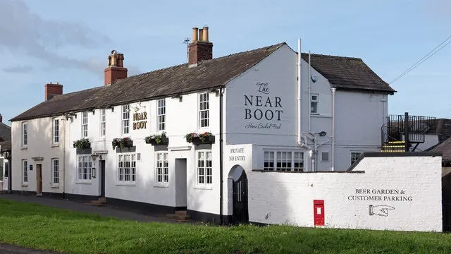 Long two-storey whitewashed pub with a slate roof and red brick chimneys, window boxes of flowers, The Near Boot painted in large black letters on the gable end, and a white wall alongside reading Beer Garden and Customer Parking with a red postbox set into it