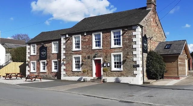 Long two-storey rubble stone pub with a slate roof, white quoins and window surrounds, a red front door and black hanging signs reading The Oak, with picnic benches on gravel to one side and a timber outbuilding to the other
