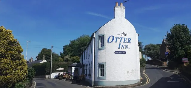 Tall whitewashed gable end with a chimney, painted in large blue letters reading the Otter Inn, Lounge and Bar, at a bend in a village road with trees and a garden