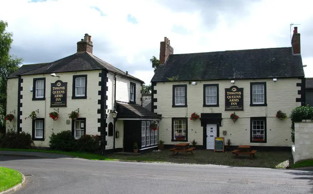 Two whitewashed two-storey buildings joined by a low porch, with slate roofs, black window frames and black corner quoins, black boards reading Thwaites Queens Arms Inn, hanging baskets and picnic benches out front